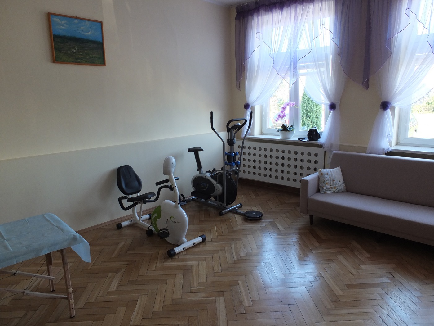Pomieszczenie do rehabilitacji w Dziennym Domu Pomocy, łóżko rehabilitacyjne, rowerek stacjonarny, orbitrek, w tle okno, po prawej stronie sofa