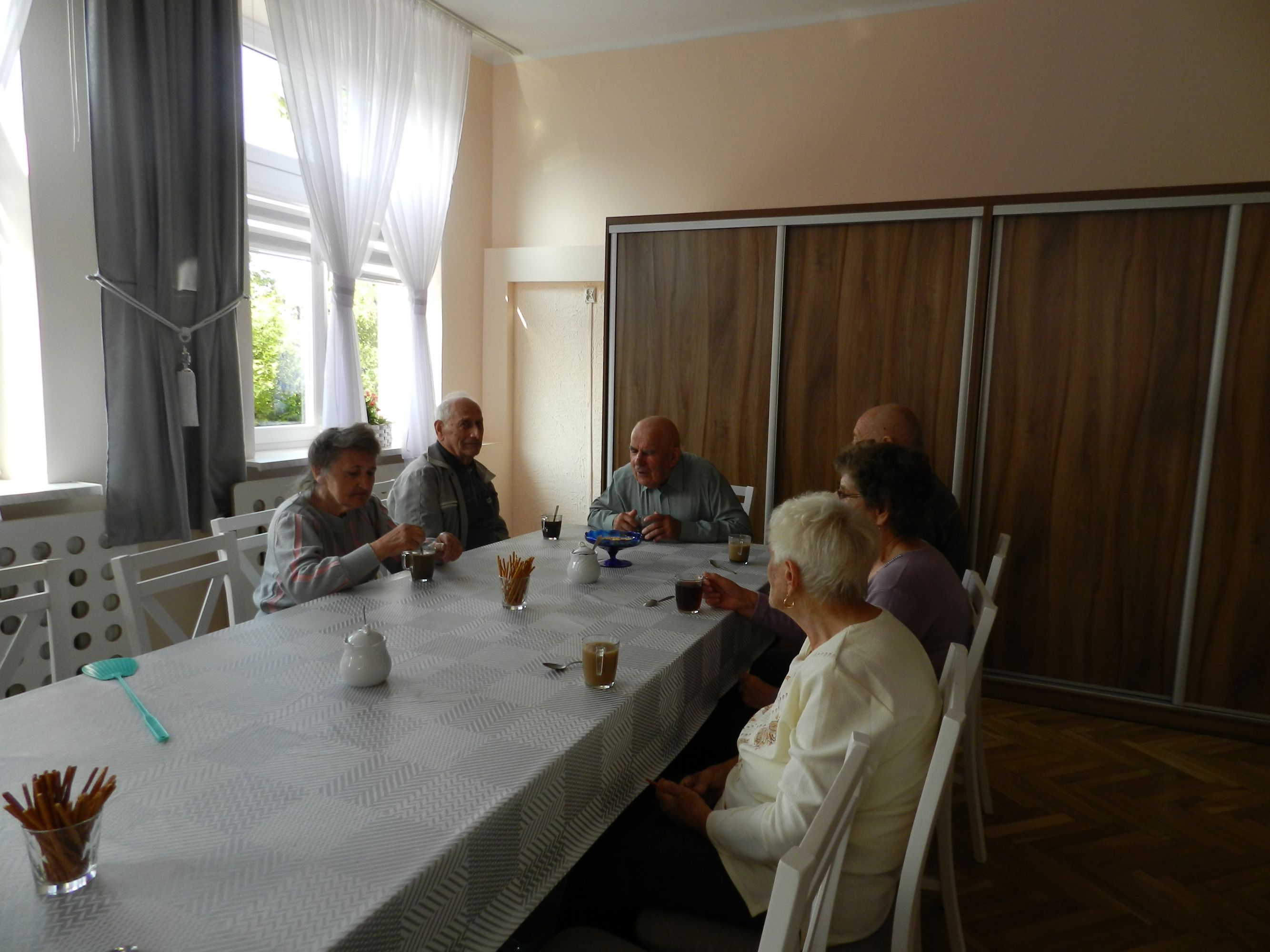 6 uczestników Dziennego Domu Pomocy przy stole podczas picia kawy, rozmowy, w tle duża prostokątna szafa, okno