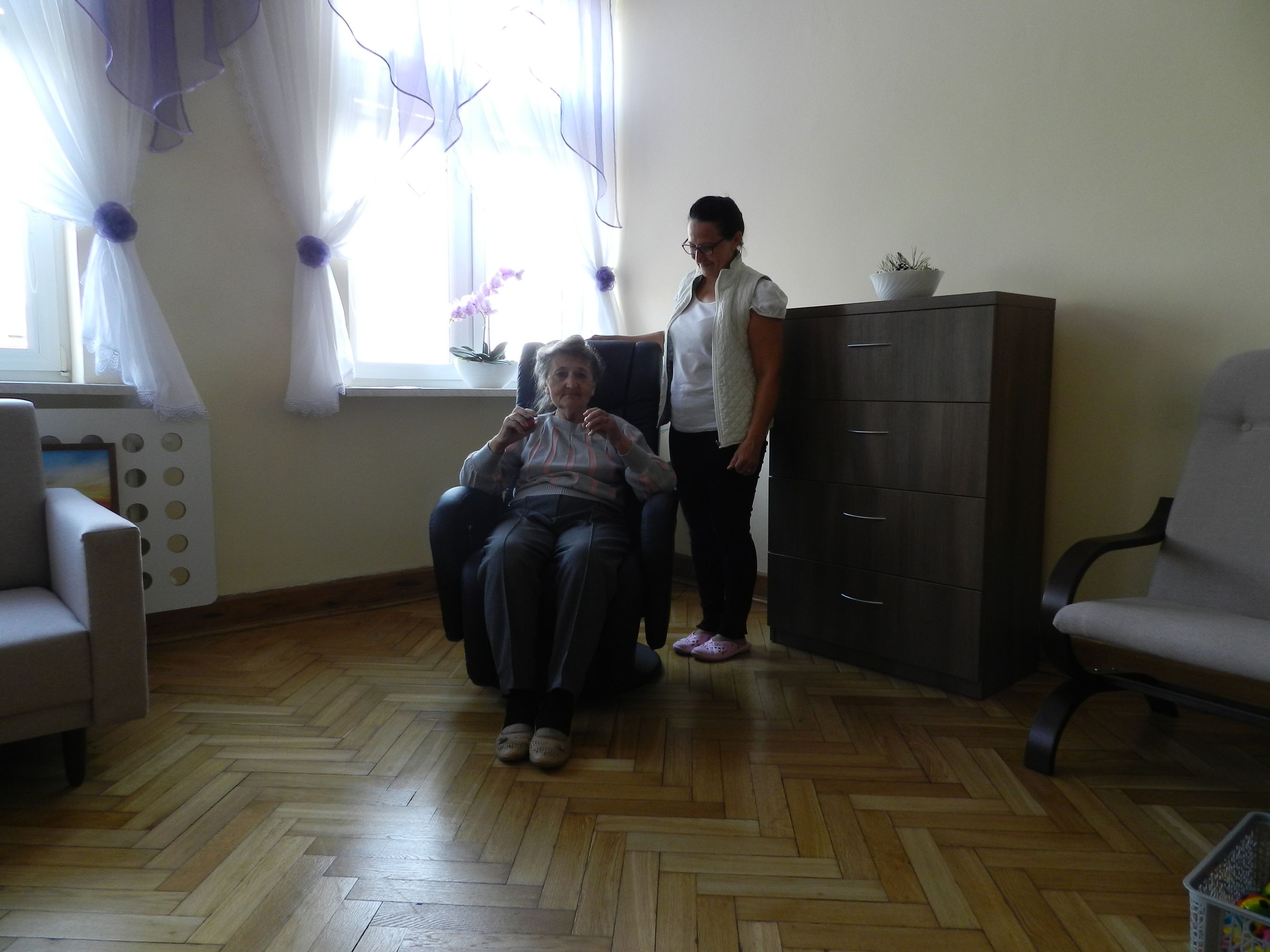 Uczestniczka Dziennego Domu Pomocy na fotelu masującym w pokoju do rehabilitacji, obok rehabilitantka, w tle okno