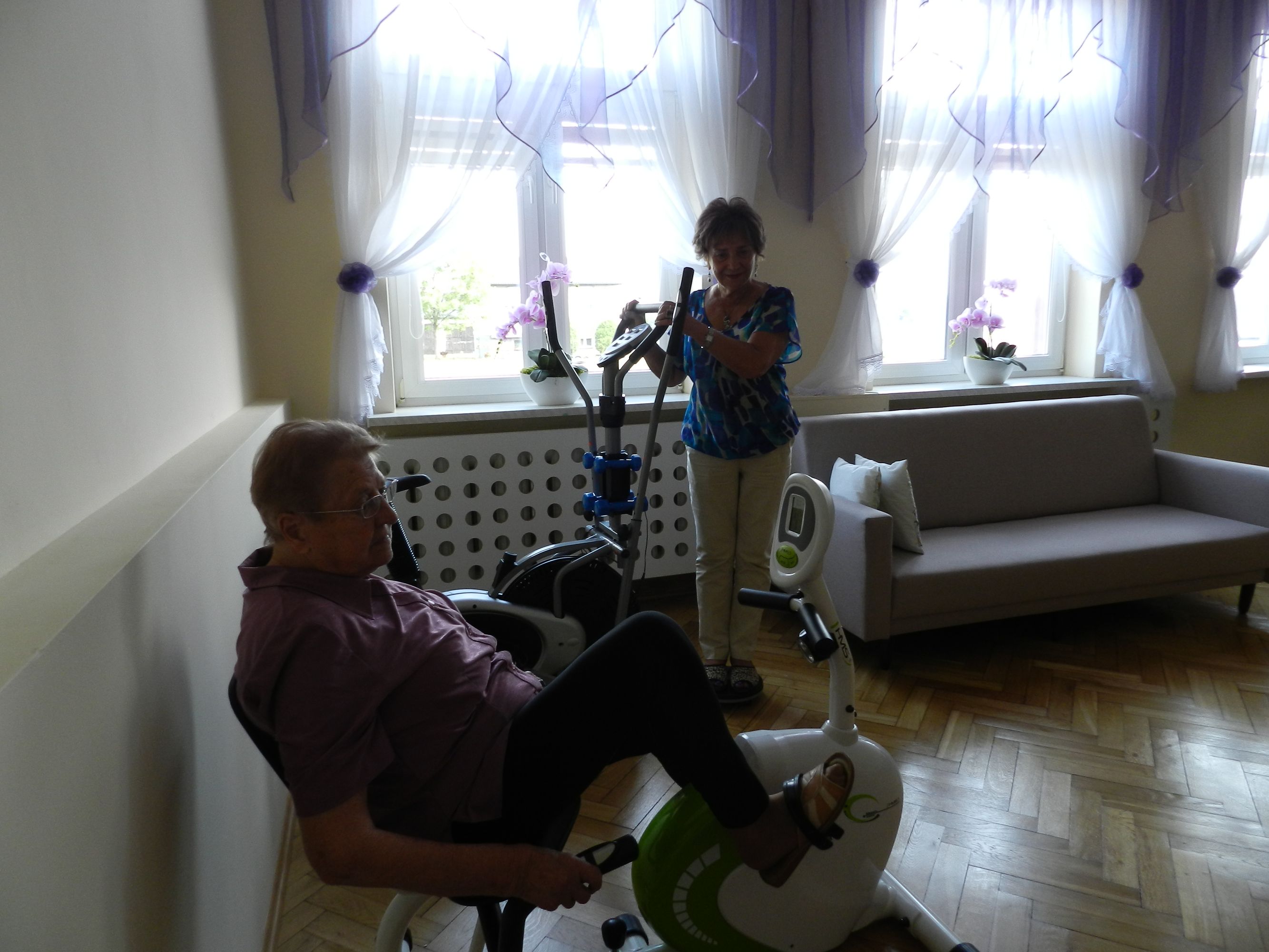 2 uczestniczków Dziennego Domu Pomocy podczas rehabilitacji na rowerku stacjonarnym i orbitreku, w tle okna