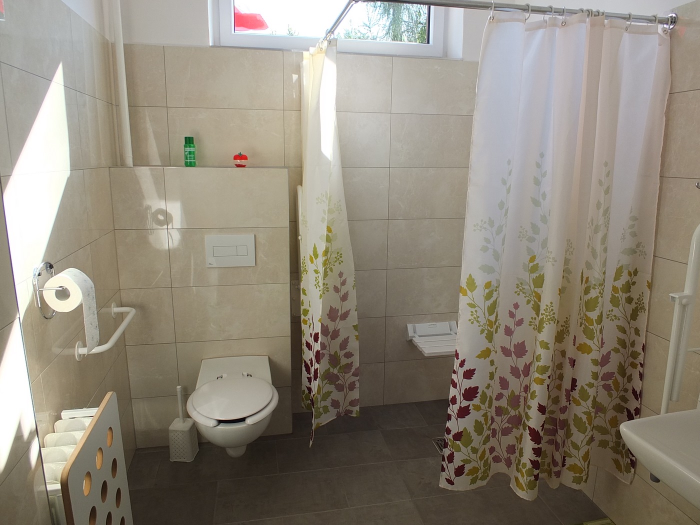 Łazienka w Dziennym Domu Pomocy dostosowana do potrzeb osób z niepełnosprawnością, brodzik niskopodłogowy, siedzisko, uchwyty, toaleta, w tle okno