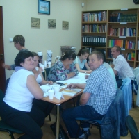Uczestnicy siedzą przy stolikach, procują w grupie przy rozłożonym na stoliku papierze.  w tle widać regał z książkami oraz powieszone na ścianie obrazki oraz fragment drzwi