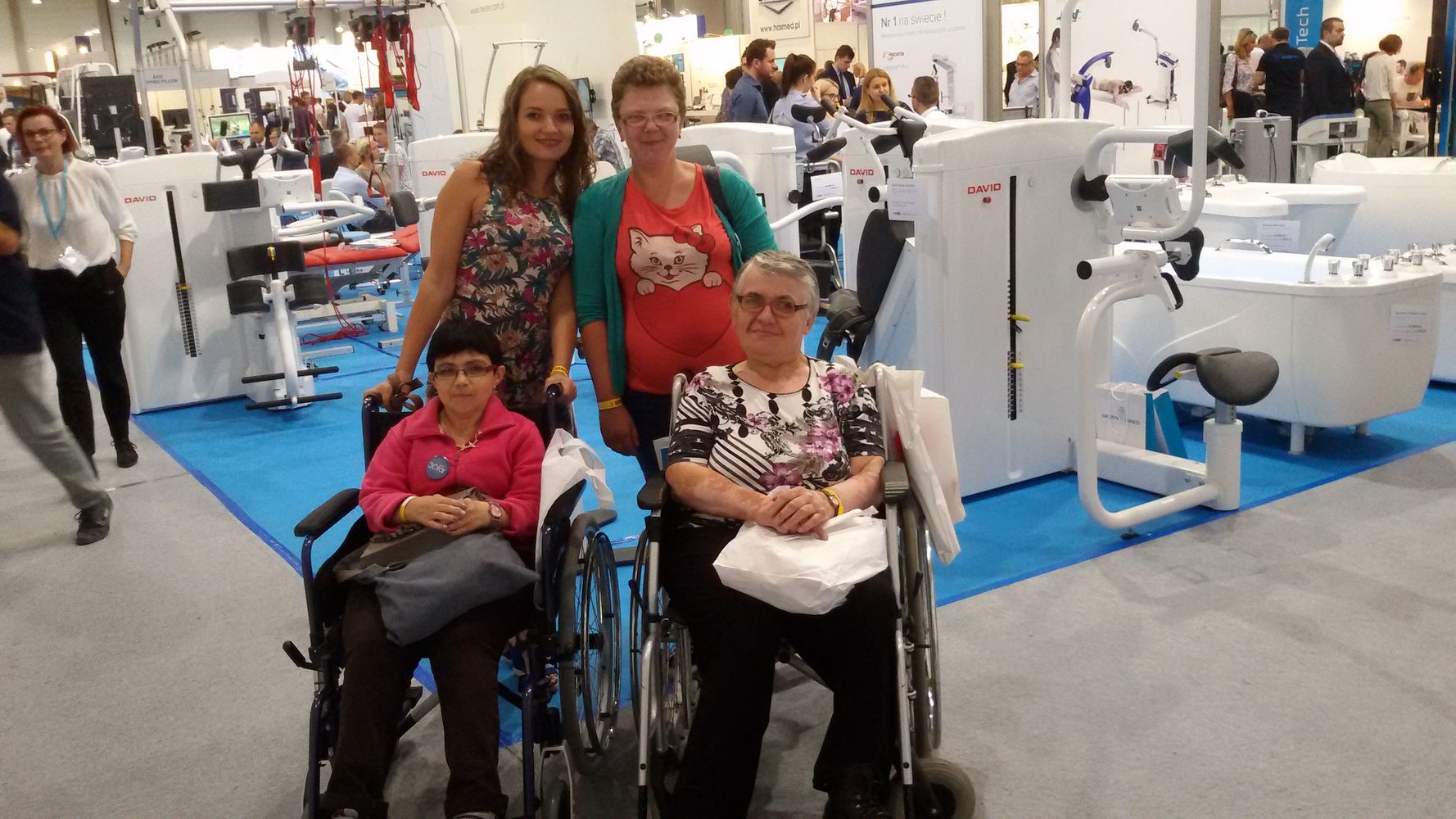 Asystent osoby niepełnosprawnej z 3 podopiecznymi - kobiety, w tym 2 kobietami na wózkach inwalidzkich podczas targów sprzętu rehabilitacyjnego.