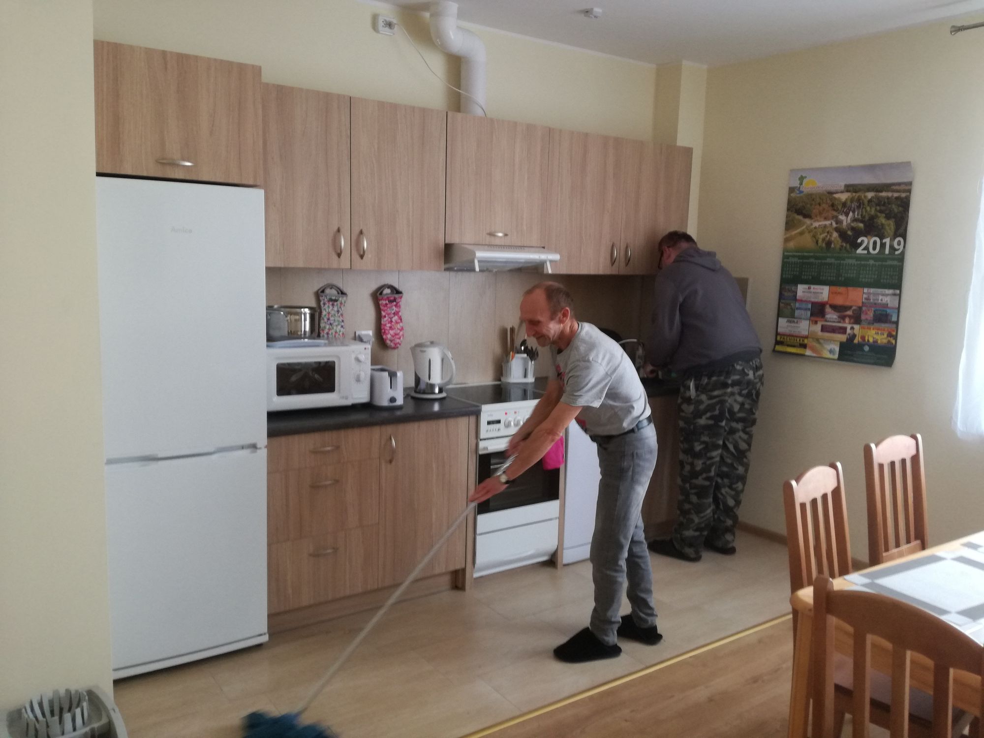 Dwaj mieszkańcy mieszkań wspomaganych podczas prac domowych, jeden zamiata podłogę w kuchni, drugi zmywa naczynia