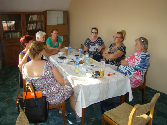 grupa 7 kobiet przy prostokątnym stole prowadzi dyskusję, w tle szafa, na stole butelki plastikowe małe z wodą